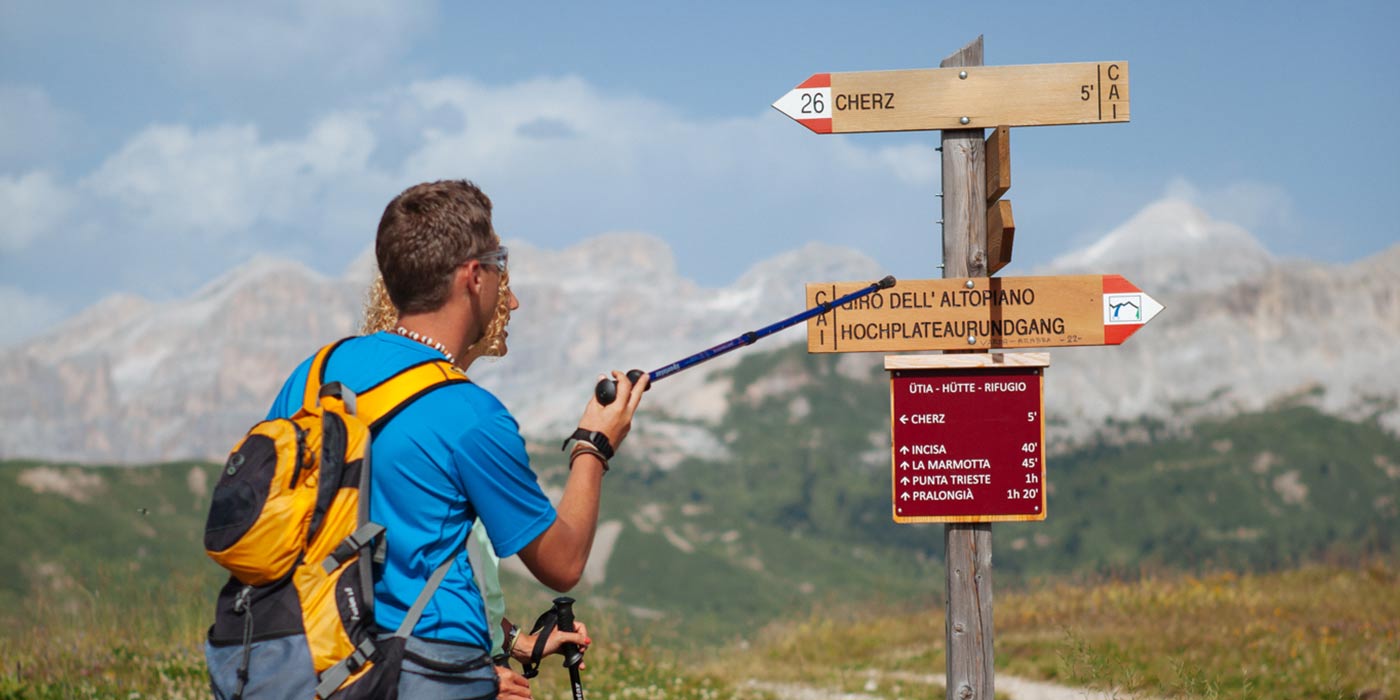 Ein Wandererpaar liest die hölzernen Wegmarkierungen mit den Dolomiten im Hintergrund