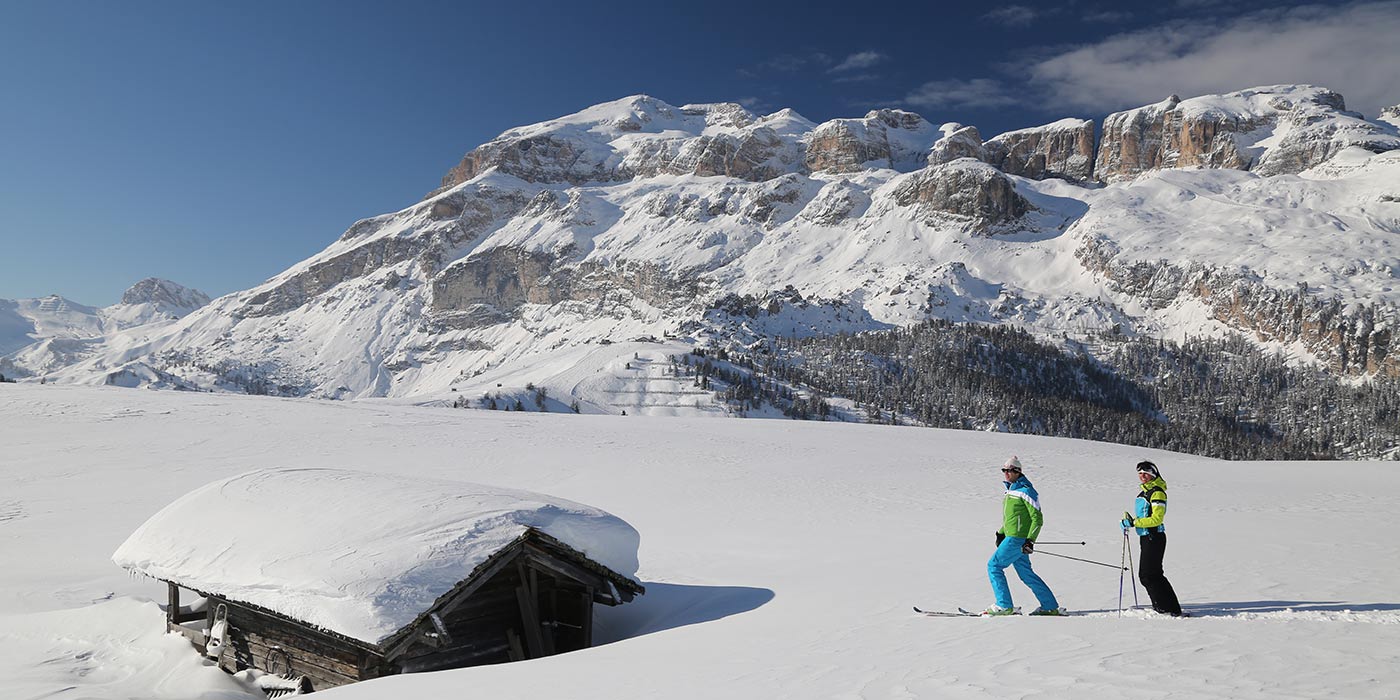 Skifahrerpaar bei einer verschneiten Hütte mit verschneitem Dolomiten-Panorama im Hintergrund