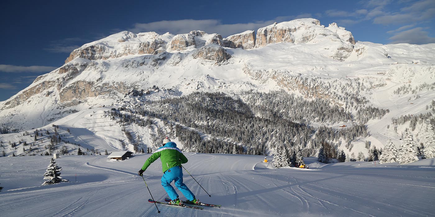 Sciatore scende lungo una pista innevata verso una baita di legno e le Dolomiti innevate