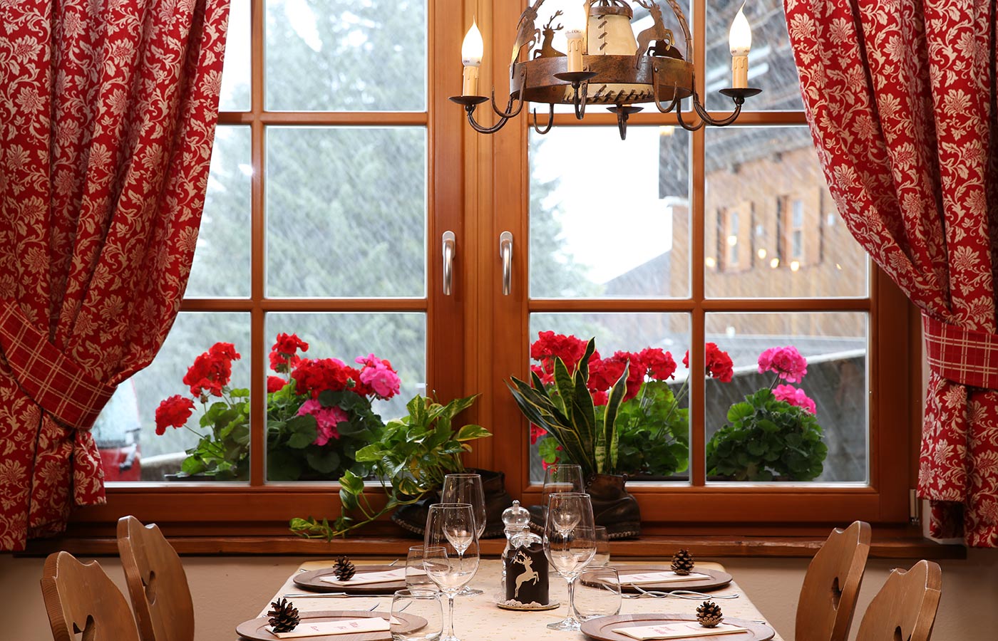 Holzfenster mit roten Vorhängen und Blumen auf dem Fensterbrett, davor ein gedeckter Tisch