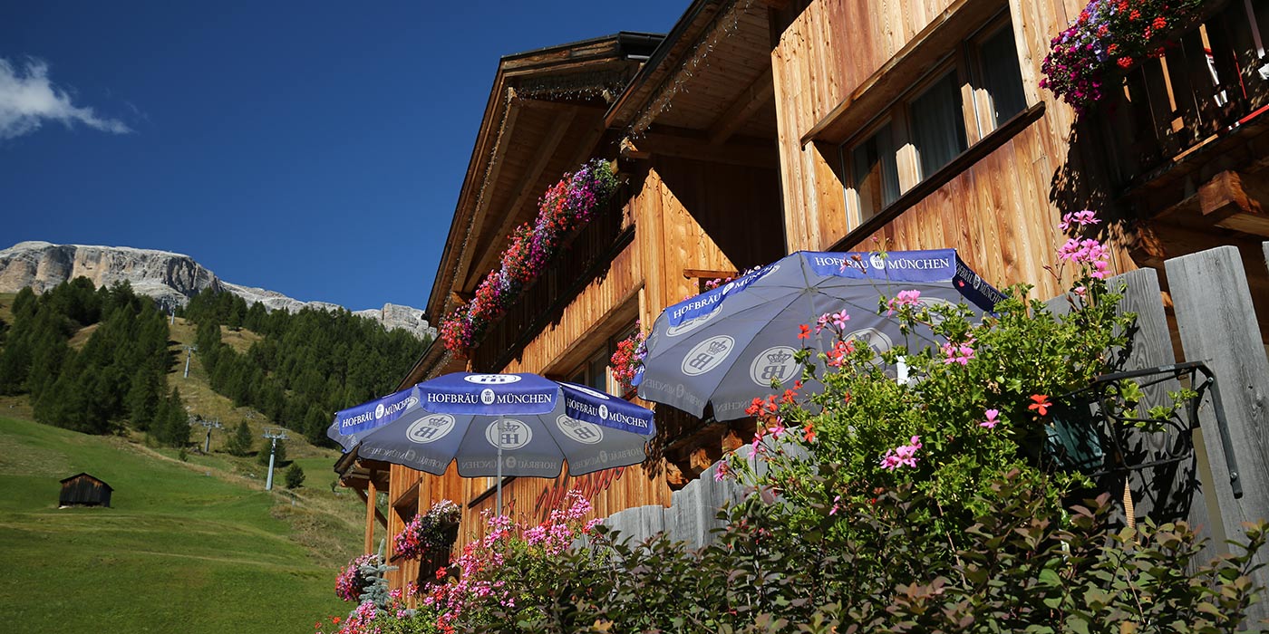 Das Hotel Mesdì im Sommer mit Sonnenschirmen und Blumen im Vordergrund, sowie den Dolomiten im Hintergrund