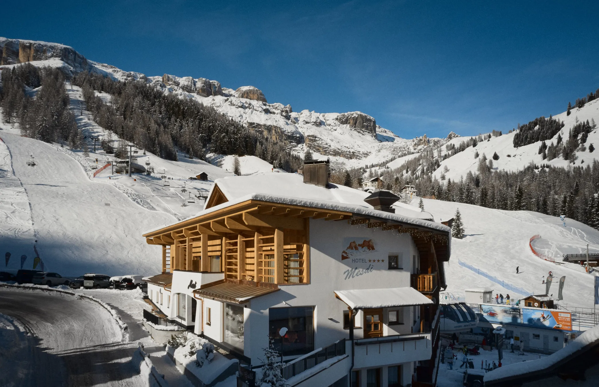Una scena alpina innevata con un accogliente chalet dal tetto di legno in primo piano. Piste da sci e montagne aspre sono visibili sotto un cielo azzurro limpido.
