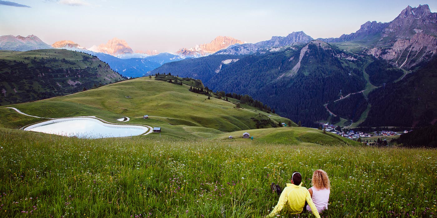 Couple sitting in a meadow near a small lake and mountain huts stares at the landscape of Arabba and the Dolomites