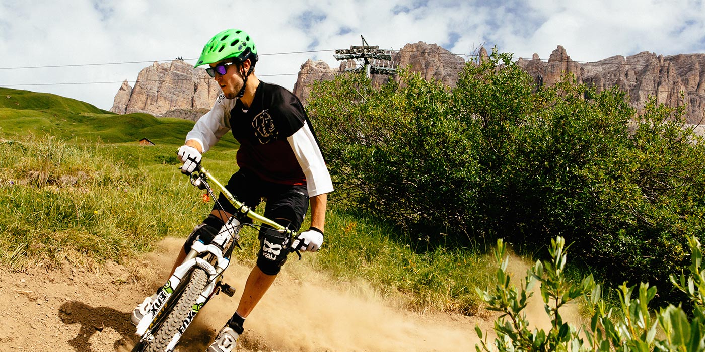 Uomo in mountain bike su una strada sterrata con prati verdi, impianti di risalita e Dolomiti sullo sfondo