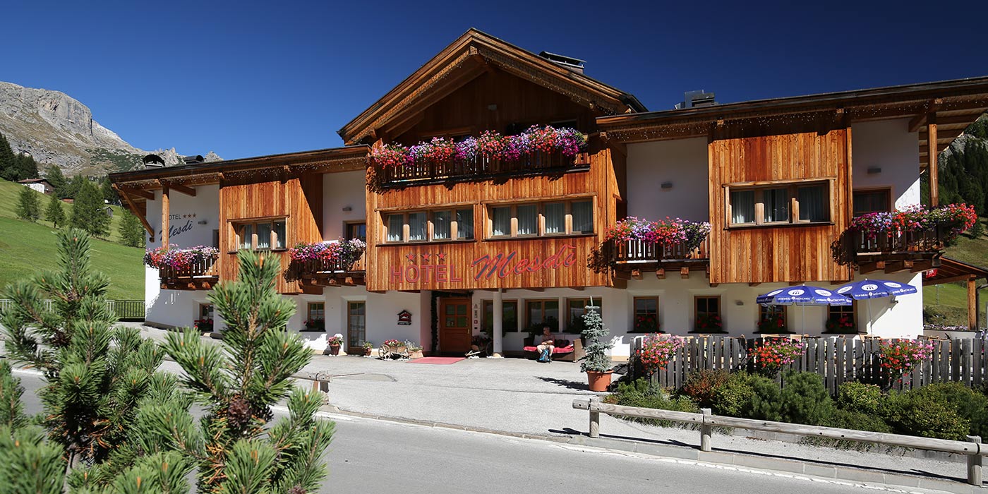 Aussenansicht des Hotels Mesdì im Sommer mit Baum im Vordergrund und Dolomiten im Hintergrund